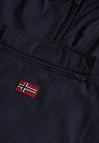 Tessuto di un giubbotto a vento blu navy con una tasca frontale con zip, dotato di un distintivo ricamato della bandiera norvegese in rosso e blu e dettagli in velcro.