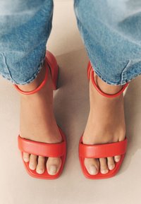Sandali rossi con tacco a blocco e cinturini alla caviglia, caratterizzati da una fascia larga per le dita e una texture in pelle liscia. Indossati con jeans blu risvoltati.