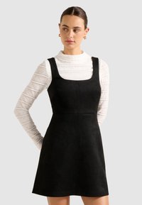 Femme aux cheveux bruns portant des boucles d'oreilles en or, un blouse en dentelle blanche à manches longues et une robe sans manches noire, regardant sur le côté.