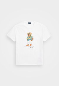 Witte katoenen t-shirt met een geborduurde beer in een kleurrijk Hawaiiaans shirt, witte broek en oranje schoenen met een zonnebril.