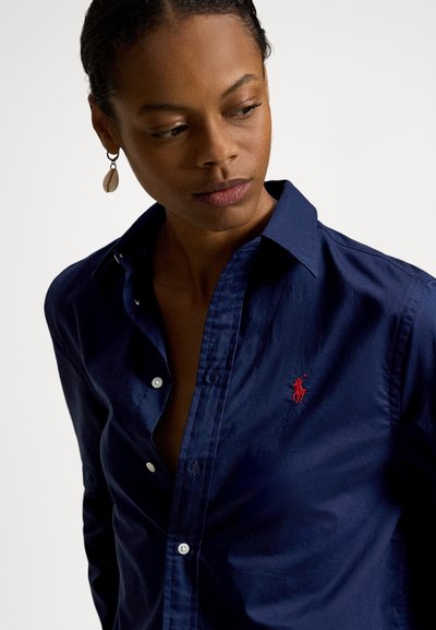 Ženska v mornarsko modri srajci z rdečim izvezenim logotipom, ki gleda navzdol in v stran, z opazno školjkino uhani.