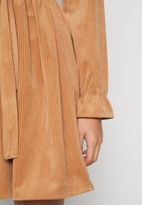 Abito in corduroy beige con righe verticali, vita cinquecchiusa e dettagli con volant sui polsini. Tessuto morbido e design sartoriale.