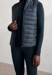 Musta puhvivesti, jossa on tikattu pinta, korkea kaulus ja vetoketjusulku istuvan pitkähihaisen paidan päällä. Saumaton muotoilu ilman lisäkoristeita.