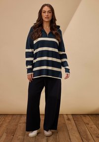 Pullover a righe blu navy e crema con colletto con zip, abbinato a pantaloni neri larghi e sneakers bianche su un pavimento di legno.