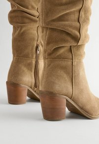 Tan suede nilkkurit, joissa on renksuyttausmuotoilu, sivuvetoketju ja pinottu puinen korko. Sileä pinta ja pyöristetty kärki.
