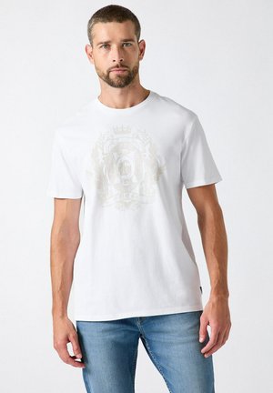 Man met kort haar en baard die een wit T-shirt met ronde hals en een vaag wapenschildontwerp draagt en een blauwe spijkerbroek staat tegen een witte achtergrond.