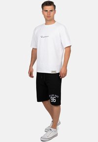 T-shirt en coton blanc avec logo en script noir, associé à un short noir avec des graphismes blancs et un cordon de serrage, complété par des baskets blanches.