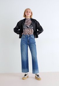 Chaqueta bomber negra, blusa de tirantes transparente a rayas, jeans de mezclilla azul de pierna ancha y zapatos con estampado. Un fondo sencillo resalta las características del atuendo.