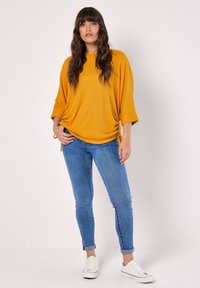 Senapsgul oversized topp med vida trekvartslånga ärmar, i kombination med figurnära blå jeans och vita sneakers. Ribbad textur; sidodragband.