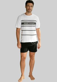 Weißes T-Shirt mit schwarzen horizontalen Streifen und dem Text "BRUNO BANANI", kombiniert mit schwarzen Shorts. Model steht barfuß vor einem grauen Hintergrund.