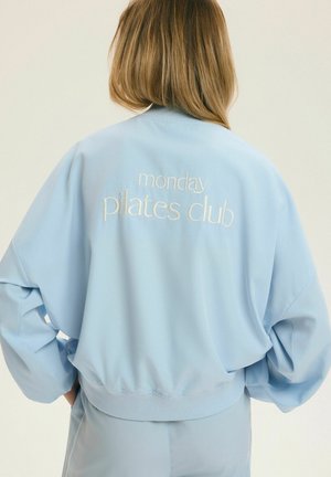 Kobieta z blond włosami do ramion, ubrana w jasnoniebieską bluzę z haftem „monday pilates club” na plecach.