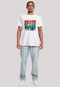 Witte katoenen t-shirt met een afbeelding van bergen en bomen in teal, oranje en roze. Gecombineerd met lichtblauwe jeans en witte sneakers.