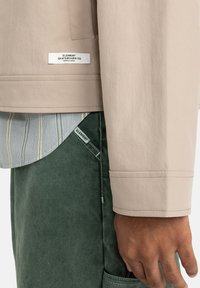 Chaqueta ligera beige con una textura suave, con una etiqueta, llevada sobre una camisa de rayas azules, combinada con pantalones verde oscuro.