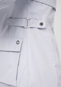 Tissu violet clair à texture lisse, équipé d'une double boucle argentée et d'un bouton pression sur une poche latérale pour une fermeture sécurisée.