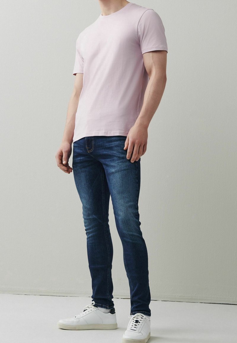 Homme portant un t-shirt rose clair à manches courtes, un jean slim bleu foncé et des baskets blanches, debout devant un mur clair uni.