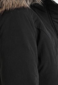 Manteau d'hiver noir avec capuche bordée de fourrure en gros plan, mettant en valeur la texture du tissu et les détails de la couture de l'épaule.