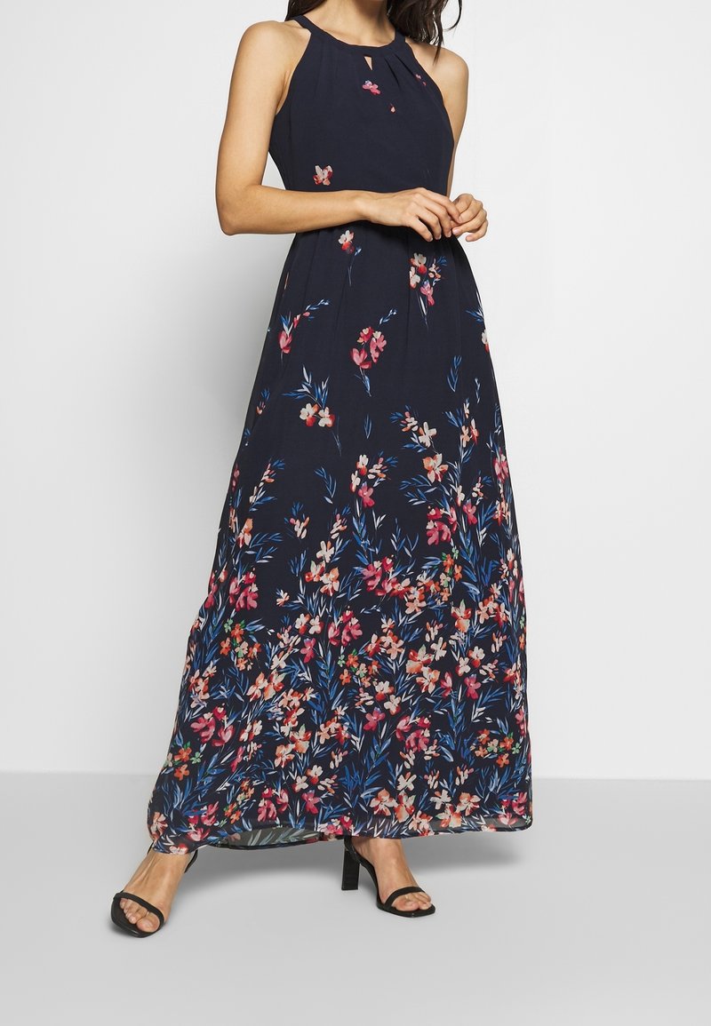 Femme portant une robe maxi sans manches bleu marine avec des motifs floraux roses et bleus, associée à des talons ouverts noirs, debout devant un fond uni.