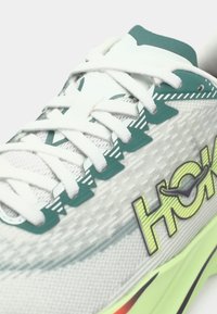Nærbillede af hvid og grøn løbesko med "Hoka"-logo på siden og detaljeret tekstureret mesh-overdel med hvide snørebånd.