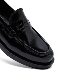 Mocasín de cuero negro brillante con una correa cosida y un detalle decorativo de corte en la parte superior, presentado sobre un fondo blanco.