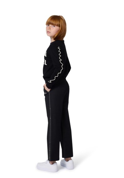 Sweatshirt noir avec lettrage blanc, associé à un pantalon noir avec une fermeture éclair sur le côté et des baskets blanches. Cheveux roux coupés au carré visibles.