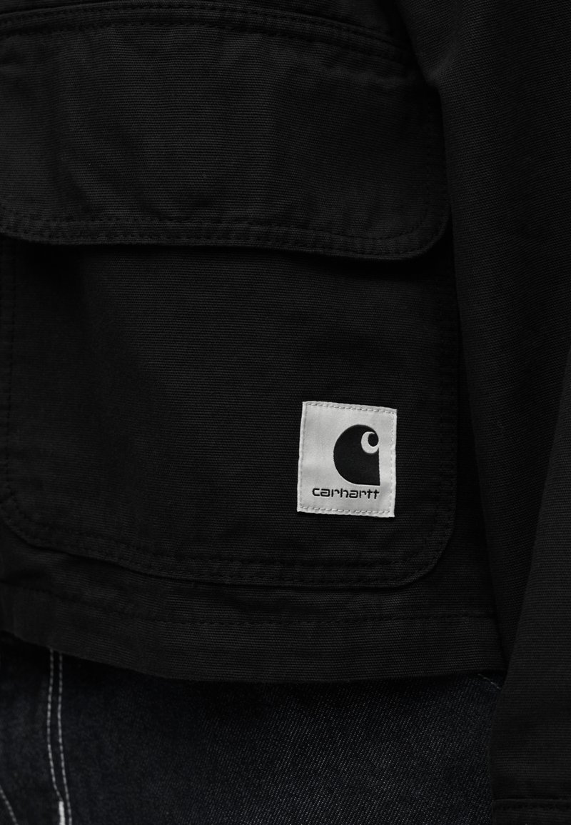 Black fabric pocket with a white square label displaying the Carhartt logo and text on clothing.
