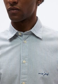 Ανοιχτό μπλε και λευκό ριγέ πουκάμισο με γιακά που κλείνει με κουμπιά, με λεπτό κεντημένο λογότυπο σε σκούρο μπλε στο στήθος.