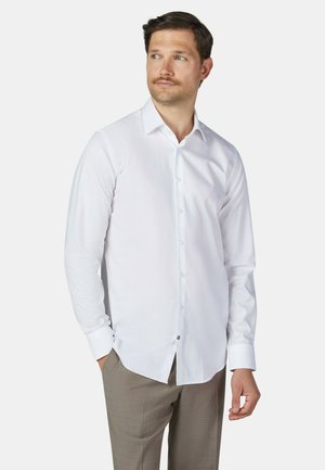 Man met snor, gekleed in een wit overhemd met lange mouwen en lichtbruine broek, staat met één hand in zijn zak en kijkt naar de zijkant.