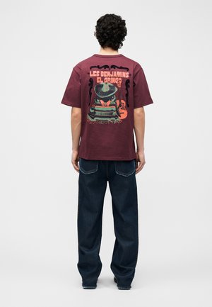 Muž s kučeravými čiernymi vlasmi v bordovom tričku s nápisom „Les Benjamins El Gringo“ a s grafikou osoby v sombrere s gitarou na chrbte.