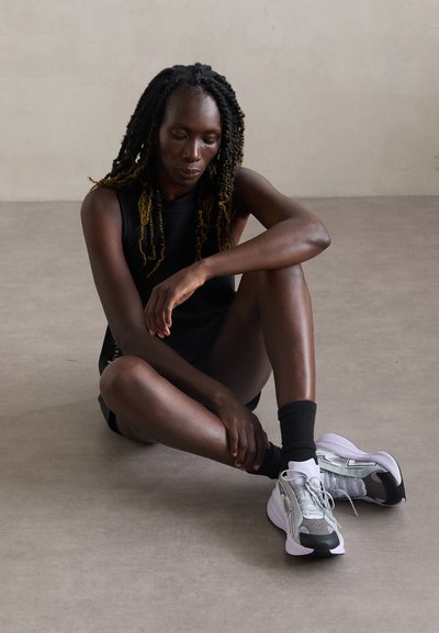 Sporty sneakers with a mixed material design, featuring shades of gray, black accents, and a textured sole. Model sits in a casual pose.