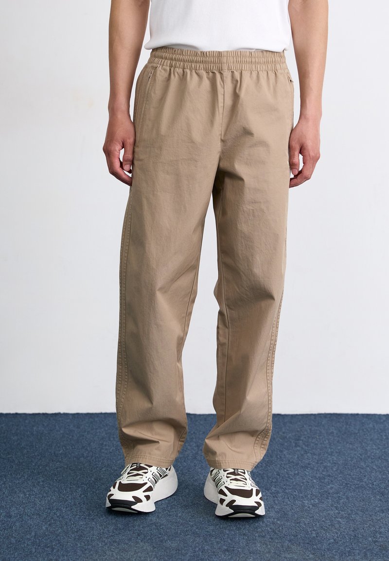 Light brown elastic waist pants with side pockets, loose fit and smooth cotton material, paired with patterned sneakers on a blue carpet.