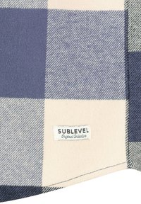 Karierte Decke mit sich abwechselnden großen navyblauen und cremefarbenen Quadraten, aus weichem Stoff gefertigt, mit einem weißen Etikett, auf dem "SUBLEVEL Original Collection" steht.