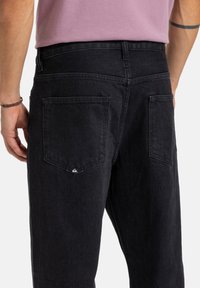 Pantaloni in denim nero visti da dietro, con due tasche posteriori decorate con un delicato logo e una texture liscia.