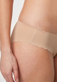 Nude brezšivne biki kopalke z valovitimi robovi, ki imajo gladek, raztegljiv material in šivano sredinsko šivko za podporo.