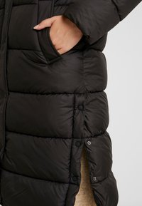 Doudoune noire avec un design matelassé, présentant une poche extérieure, des boutons-pression à l'ourlet et une finition lisse et brillante.
