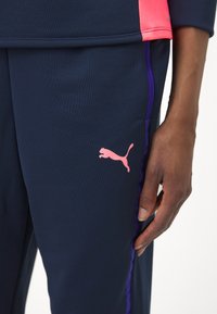 Puma sportska odjeća s tamnoplavom tkaninom, ruži�častim detaljima i istaknutim logotipom. Uključuje ljubičaste bočne pruge i teksturirani dizajn.