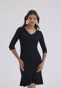 Czarna sukienka w turkusowe kropki, dekolt w serek, rękawy trzyczwarte i falbaniasty dół. W zestawie duże, złote owalne kolczyki. Gładka tekstura.