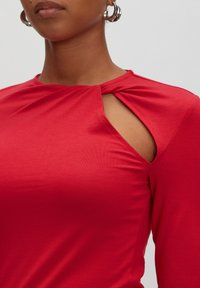 Rotes Langarmoberteil aus glattem Stoff, mit einer Twist-Detail nahe dem Ausschnitt und einem angesagten Ausschnitt an der linken Schulter.