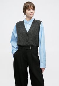 Pelēks adīts vests ar V-veida izgriezumu, pār kārtots gaiši zilas svītrains krekls, kombinēts ar melnām plašām biksēm ar pogu rotājumiem.