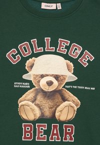 Zaļais džemperis ar grafiku, kurā attēlots teddy lācis, kas valkā spaiņu cepuri. Uzraksts saka "KOLLEĢU LĀCIS" ar papildu frāzēm baltā un bordo krāsā.