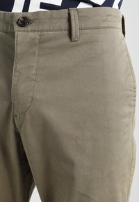 Pantaloni di cotone verde oliva con una texture liscia, chiusura a bottone e tasche posteriori. Cuciture dettagliate lungo le cuciture e la vita.