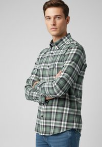 Camisa de franela a cuadros verdes con acentos en negro y blanco, con botones en la parte delantera, cuello y bolsillo en el pecho, combinada con pantalones vaqueros azules.