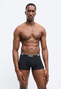 Homme musclé debout portant un boxer BOSS noir sur un fond clair uni, face à l'avant avec une expression neutre.