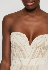 Hüftfreies Kleid mit tiefem V-Ausschnitt, strukturiertem beigem Stoff mit goldenen geometrischen Mustern und einem stabilen Oberteil für Unterstützung.