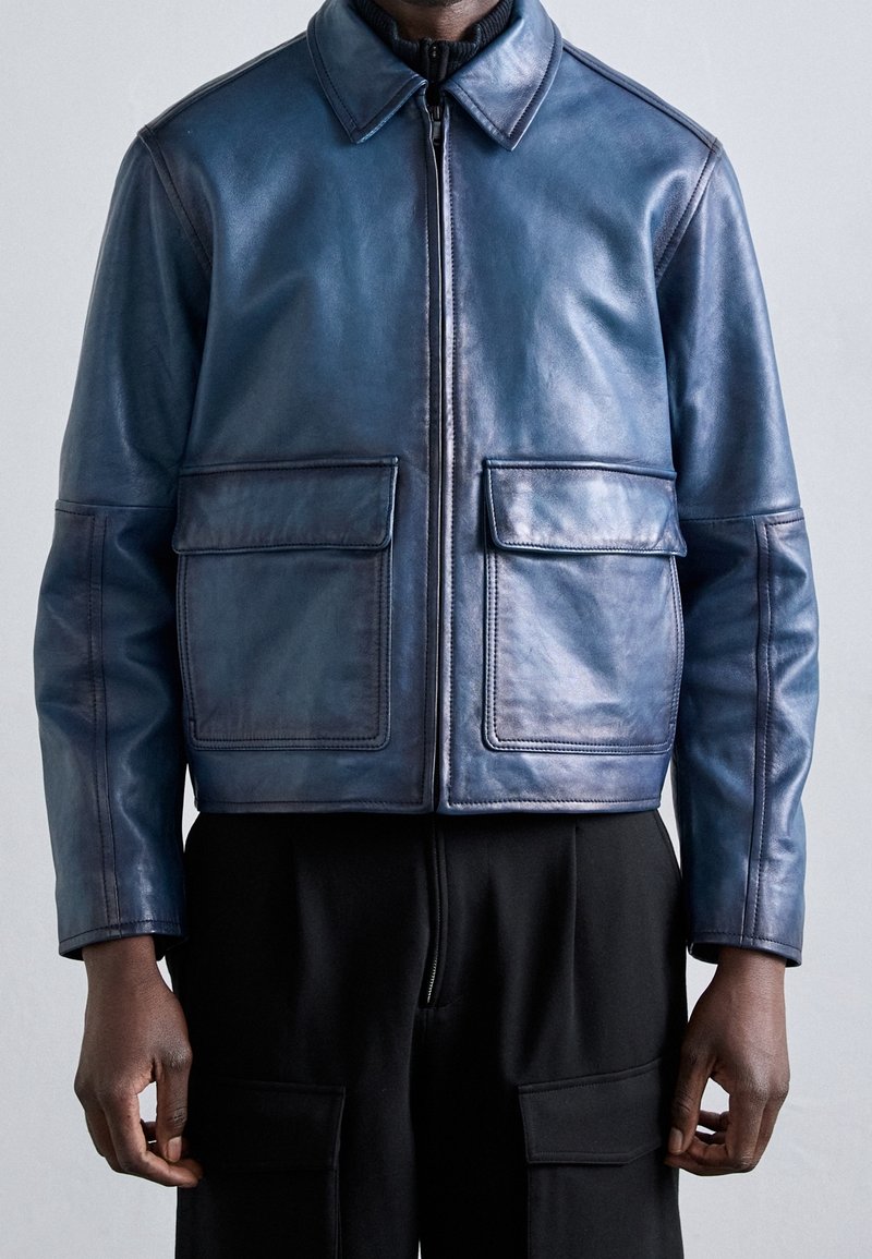 Mężczyzna ubrany w niebieską skórzaną kurtkę na zamek z dużymi przednimi kieszeniami z patkami oraz czarne spodnie z kieszeniami typu cargo.