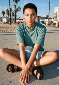 Jeune personne aux cheveux courts et bouclés assise en tailleur sur un béton près de la plage, portant un t-shirt turquoise, un short en jean et des sandales noires.