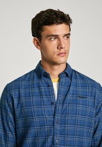 Camicia a quadri blu realizzata in tessuto morbido, con bottoni, colletto e accenti gialli nel motivo a quadri. Indossata sopra una camicia chiara.