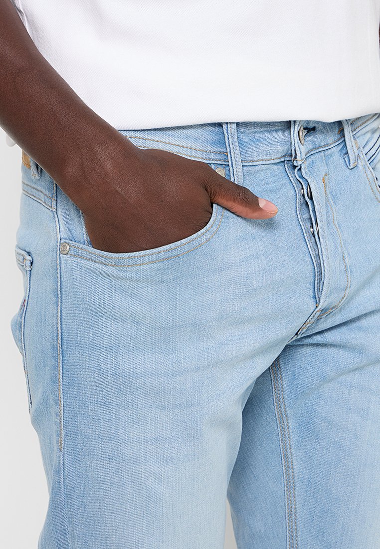 Lichtblauwe denimjeans hebben een slim fit, met een zichtbare voorzak en metalen knoophardware. De stof heeft een zachte, iets gestructureerde uitstraling.