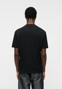 Άνδρας που στέκεται γυρισμένος προς τα πίσω, φορώντας μαύρο απλό κοντομάνικο μπλουζάκι και σκούρο τζιν παντελόνι, με φόντο απλό λευκό.