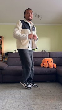 Chaqueta de forro polar blanca y negra, pantalones grises y zapatillas negras. Un pulpo de peluche naranja sobre un sofá marrón oscuro en una habitación bien iluminada.
