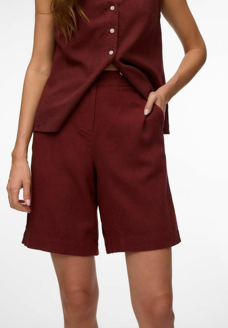 Femme portant un haut sans manches bordeaux boutonné et un short assorti jusqu'au genou, une main dans la poche, sur fond blanc.